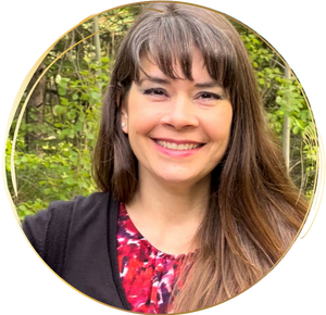 Circle picture with gold around it and Kristie centered smiling, straight long, medium brown hair wearing a red print shirt and a black v-neck cardigan sweater. There are green blurred deciduous trees in the background.